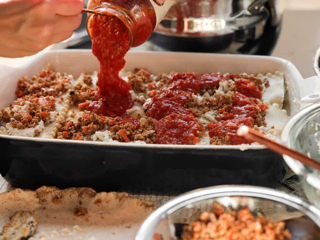 A person pours tomato sauce over a vegan lasagna in a black baking dish. The dish is layered with pasta sheets, cooked ground vegan italian sausage, and sauce. Other ingredients and utensils are visible on the surrounding countertop.